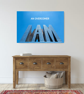 A canvas print with the text 'AN OVERCOMER I AM' in bold, stylized lettering on a blue background, hung above a wooden console table.