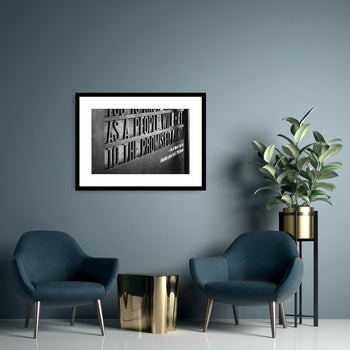 A framed inspirational art piece with text, displayed on a wall between two armchairs and a side table with a plant.