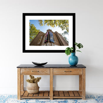 A framed artwork of the Carnegie Visual Arts Center in Decatur, Alabama, displayed on a wall above a wooden shelf with a decorative plant and a vase.
