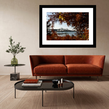 A framed color photo of the Tennessee River Bridge in Decatur, Alabama, displayed on a wall above a brown leather sofa with a wooden flooring and minimalist decor.