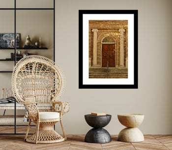 A framed artwork featuring a doorway with a classical design, displayed on a wall above a contemporary wicker sofa and a modern wooden side table in a well-lit, elegant living space.