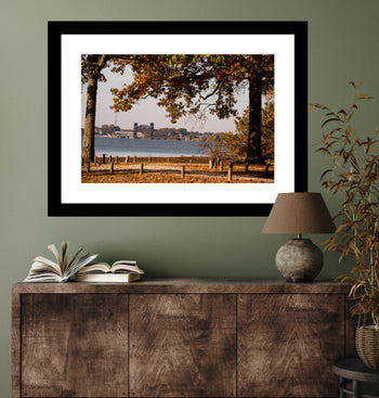 A framed art print featuring an old railroad bridge in Decatur Alabama, with fall trees in the foreground. The print is displayed on a wall above a wooden cabinet with a lamp and books.
