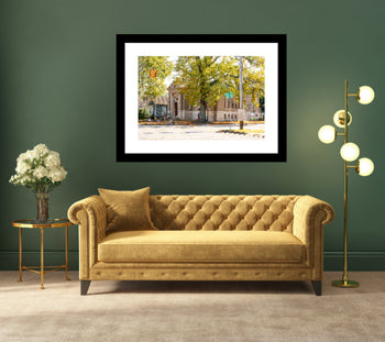 A framed artwork featuring a photo of a street corner in Decatur, Alabama, with a building on the corner, trees, and a car. The artwork is displayed above a yellow sofa in a room with green walls and a gold floor lamp.