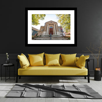 A framed artwork featuring the front view of the Carnegie Visual Arts Center building in Decatur, Alabama, displayed on a wall above a yellow sofa with black pillows.