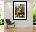 A framed artwork featuring a photograph of Bank Street Green Private Park in Decatur, Alabama, displayed on a wall above a modern chair in a bright room with windows overlooking the park.
