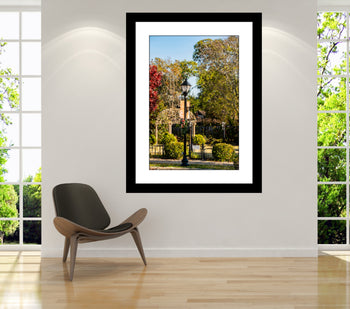 A framed artwork featuring a photograph of Bank Street Green Private Park in Decatur, Alabama, displayed on a wall above a modern chair in a bright room with windows overlooking the park.