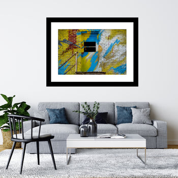 A framed artwork featuring a colorful abstract painting of an abandoned building in Decatur, Alabama, displayed on a wall above a sofa with minimalist furniture in a living room setting.