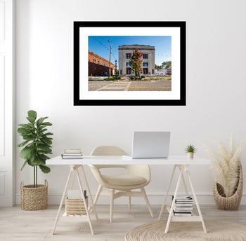 A framed artwork of the Veterans Building, Downtown Decatur, Alabama, displayed on a wall above a desk with a laptop and plant.