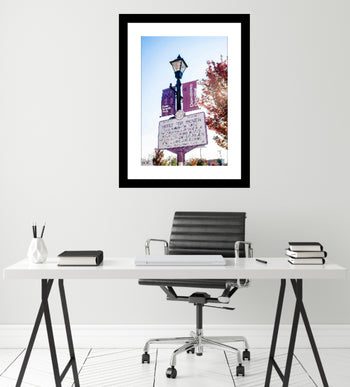 A framed artwork featuring a 'Votes For Women' sign in Decatur, Alabama, displayed on a wall above a desk with a lamp and books.