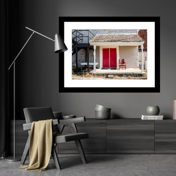 A framed artwork featuring a red door and a white home, with a red chair and a lamp in the foreground, displayed on a wall in a modern living room setting.