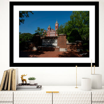 A framed canvas print featuring Auburn University's Samford Hall with a clear blue sky in the background. The frame is white, and the artwork is displayed on a shelf with books and decorative items around it.