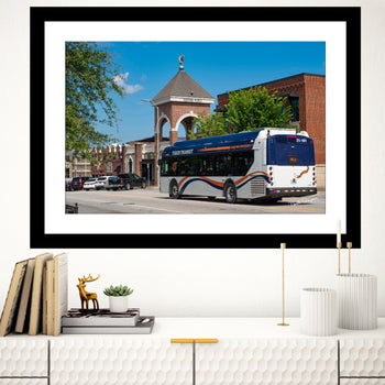 A framed art print of a bus on a street corner with a building in the background, displayed on a wall above a bookshelf with decorative items.