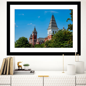A framed art print featuring a photograph of Hargis Hall and Samford Hall on the Auburn University campus, displayed on a wall above a bookshelf with decorative items.