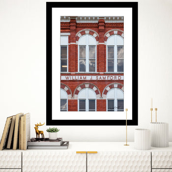 A framed art print featuring a photograph of Samford Hall with the text 'WILLIAM J. SAMFORD' on the front, displayed on a wall above a bookshelf with books and decorative items.