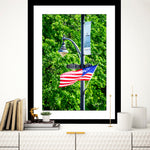 A framed artwork featuring an American flag on a street corner in Guntersville, Alabama, displayed on a wall in an interior setting with books and decorative items around it.