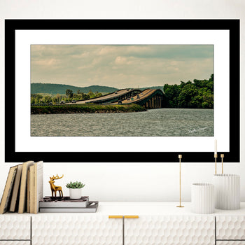 A framed artwork featuring a photograph of the Guntersville Bridge over water, displayed on a wall in a home setting with books, a deer figurine, and candle holders beside it.