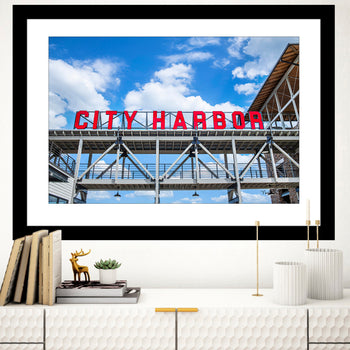 A framed wall art print featuring the text 'City Harbor' on a sign with a blue sky and clouds in the background, displayed in a home setting.