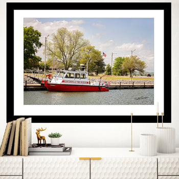 A framed artwork featuring a photograph of a red and white fire rescue boat on water, with a clear blue sky and trees in the background, displayed on a white wall in a home setting.