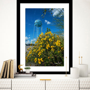 A framed artwork featuring a water tower with yellow flowers in the foreground, displayed on a wall above a bookshelf with books and decorative items.