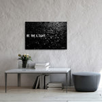 A canvas print with the phrase 'BE THE LIGHT' displayed in black and white, hung on a wall above a table with books and a vase of flowers.