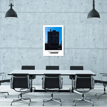 A framed inspirational artwork featuring a building at dusk, hanging on the wall in a modern office setting with concrete walls and fluorescent lighting.