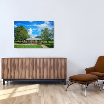 A framed inspirational art piece depicting a old house set against a blue sky with trees, displayed on a wall above a wooden cabinet and a modern chair.