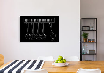 A canvas print featuring Newton's cradle with the text 'Positive Energy Only Please' displayed at the top, positioned on a wall above a table in a room with two chairs and a bookshelf.