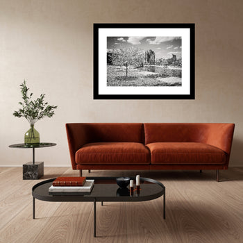 A black and white photo of cherry blossom trees in a park, framed in a black frame, displayed on a wall above a brown sofa and a transparent coffee table, with a vase of flowers and a book on the table.