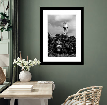 A black and white photo of a water tower with a 'STOP' sign in front, framed and displayed on a wall above a wooden table and wicker chair in a room with a green paneled wall.