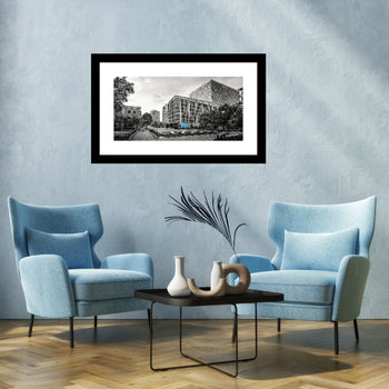 A black and white photograph featuring a panoramic view of Big Spring Park with historical buildings in the background, framed in a white frame, displayed on a wall above a blue sofa and a black table.