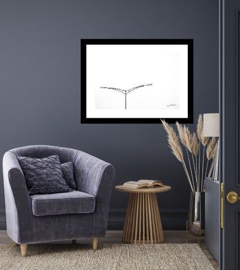 A black and white photograph of birds on a light post, framed in a simple black frame, displayed on a wall above a grey chair with a cushion, next to a round wooden side table with a lamp and a decorative plant.