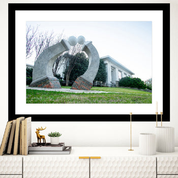A framed art canvas print featuring a sculpture of two hands holding each other in front of the Huntsville Museum of Art, with a building and trees in the background.