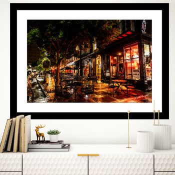 A framed art print featuring an outdoor seating area of a restaurant with illuminated tables and a tree in the background, displayed on a white wall in an interior setting.