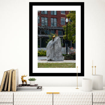 A black frame fine art print featuring a photograph of a natural rock formation in a city park, with buildings and trees in the background.