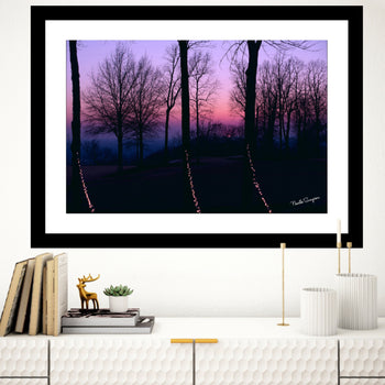 A framed artwork featuring a silhouette of trees against a purple sunset sky, displayed on a wall above a bookshelf with a decorative deer figure.