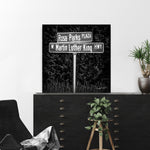 A framed poster of a street sign at the intersection of Rosa Parks Way and Martin Luther King Jr. Blvd, with a black and white color scheme, displayed on a wall above a wooden cabinet with a potted plant on it.