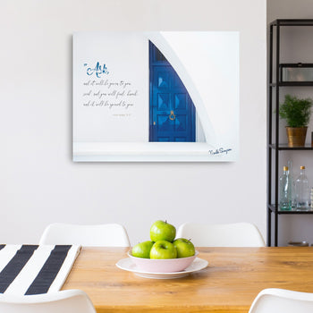 A framed canvas print featuring a blue door with text overlay, displayed on a wall above a table with green apples and magazines.