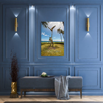 A framed picture featuring a beach scene with palm trees and a figure standing in the foreground, displayed on a blue wall above a bench with a grey cushion and a gold decorative object to the left.