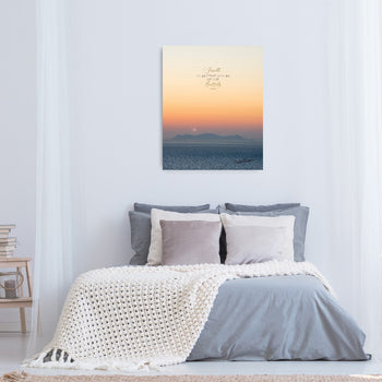A framed canvas print with a sunset over a body of water, hung on a white wall above a bed with grey bedding and a beige blanket.