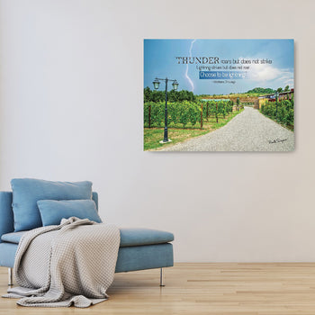 A framed canvas print with a quote displayed on a wall, depicting a landscape of Tuscany, Italy, with a road and greenery, placed above a blue sofa with a beige blanket.