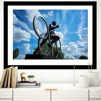 A framed artwork featuring a bronze statue of Jesse Owens, set against a blue sky with clouds, displayed in a home interior setting.