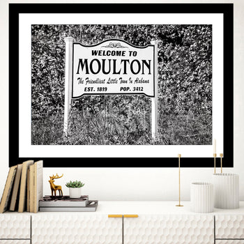 A black and white photo of a 'Welcome to Moulton' sign, displayed as a fine art print without a mat or frame, positioned on a wall above a shelf with books and decorative items.