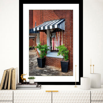 A framed photo of a building facade featuring an American flag, striped awning, and potted plants, presented in a home decor setting.
