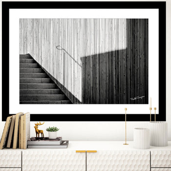 A black and white photo art print featuring a staircase with sunlight and shadows, displayed on a wall above a minimalist bookshelf with a plant and decorative items.