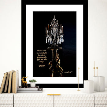 A framed art print featuring a black and white photo of a candlestick holder with a quote, displayed on a wall above a bookshelf with a plant and decorative items.