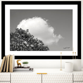 A black and white photo of a big cloud in the sky behind some trees, displayed as a wall art above a minimalist bookshelf with books and decorative items.